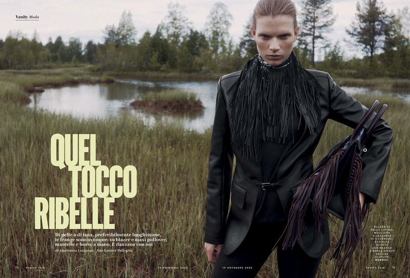 Fashion editorial shot featuring a model in a black leather jacket and fringed scarf, posing in a grassy field with a lake and trees.