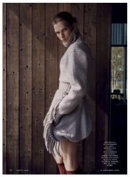 Model Sara Eirud wearing a chunky high-neck grey knit sweater dress with fringe detailing against a vertical wood plank wall.