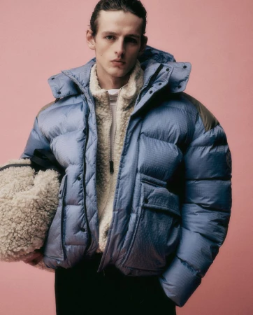 Portrait of a young adult male wearing a light blue puffer jacket over a sherpa fleece jacket, standing in front of a pink background.