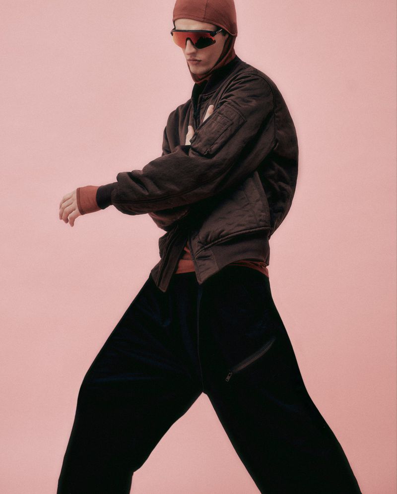 Male model wearing a brown bomber jacket, black pants, and a head covering, striking a pose against a light pink backdrop.