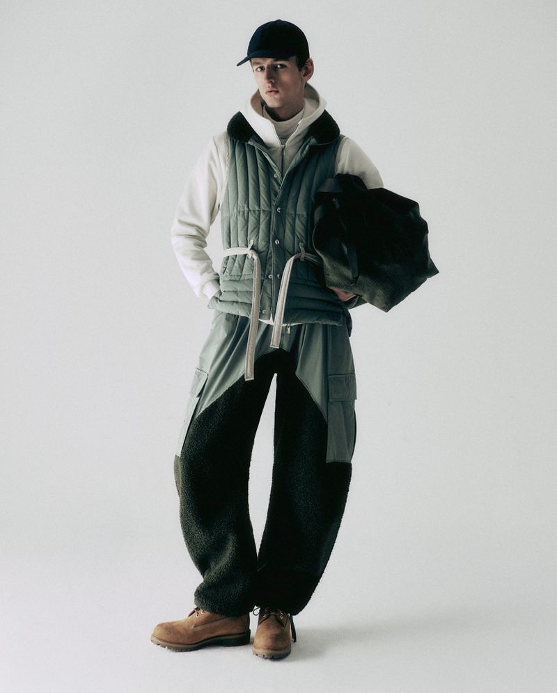Male model wears a cap, quilted vest, hoodie, two-tone pants, and work boots in a studio fashion shot on a seamless white backdrop.