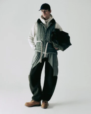 Male model wears a cap, quilted vest, hoodie, two-tone pants, and work boots in a studio fashion shot on a seamless white backdrop.