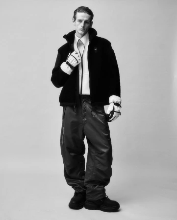 Monochrome portrait of a man in a black sherpa jacket, white shirt, grey pants, and black shoes, wearing white ski gloves in a studio.