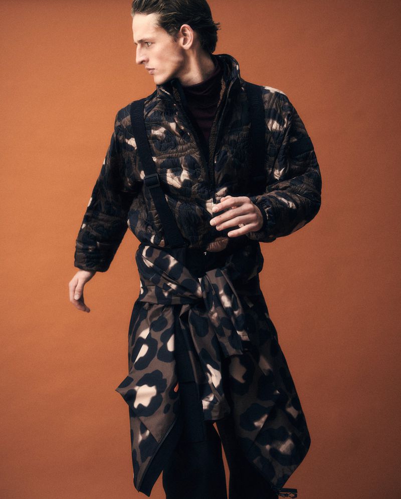 A male model poses in a patterned jacket and pants, tied with a scarf, against a bronze backdrop in a fashion studio shot.