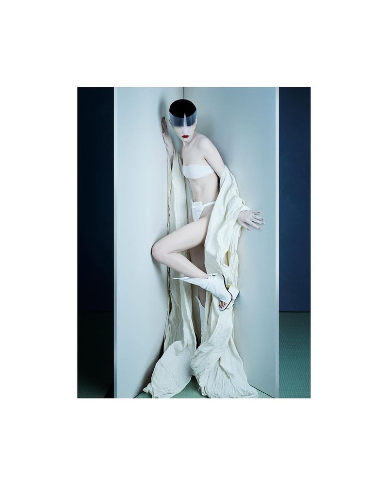 Fashion photography: A slender woman in a white lingerie-style outfit with avant-garde visor and sculptural sandals poses in a corner.