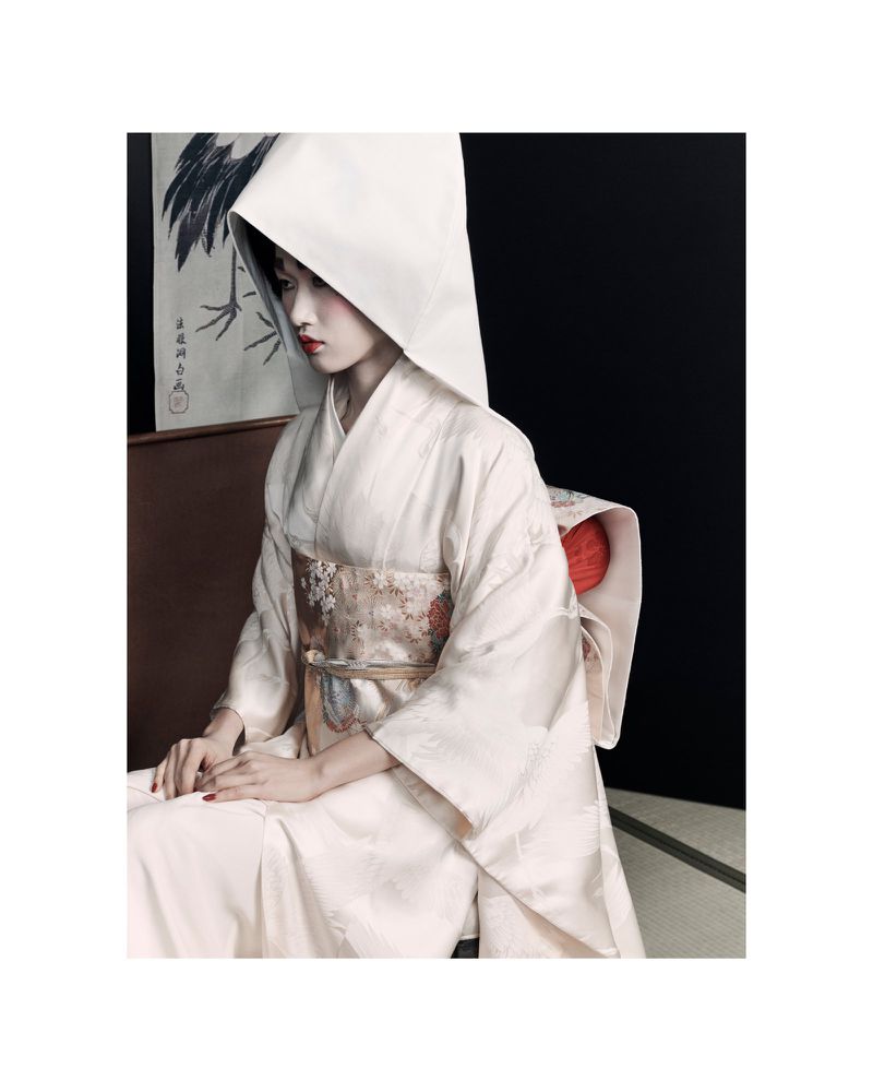 Asian woman in traditional white kimono and headdress sits indoors against a black backdrop with a crane painting in soft light.