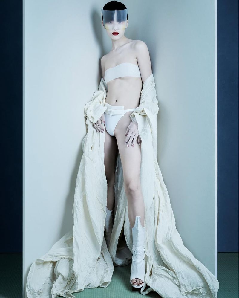 Avant-garde fashion shot of a slender adult female in a white outfit, visor, and boots, leaning against an architectural corner in studio lighting.