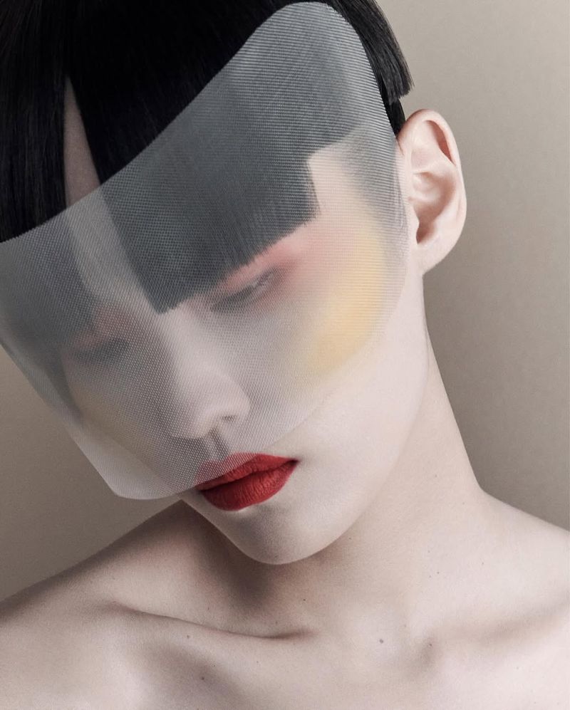 Close-up portrait of an adult woman with pale skin, red lipstick, and dark hair, partially obscured by a semi-transparent mesh face covering.