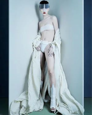 Model in a white bandeau and voluminous draped cream coat with a translucent visor standing in a studio corner.