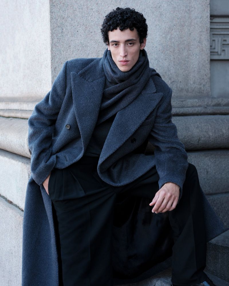 Portrait of a young adult male model sitting on stone steps wearing a gray overcoat and scarf, looking directly at the camera.