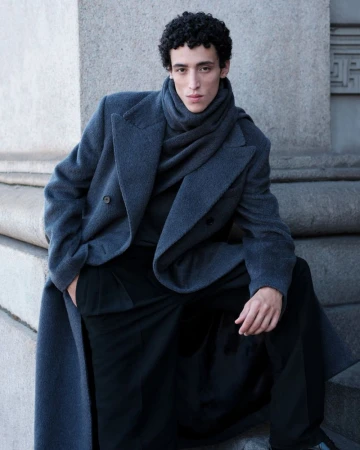 Portrait of a young adult male model sitting on stone steps wearing a gray overcoat and scarf, looking directly at the camera.