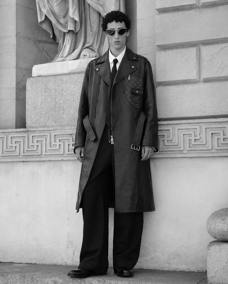 Monochrome portrait of a stylish male model in sunglasses and trench coat, posing outdoors against an ornate architectural backdrop.