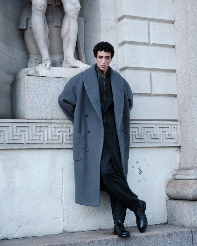 A fashionable young adult male with curly hair poses in an oversized blue-grey coat leaning against a grey stone architectural wall.