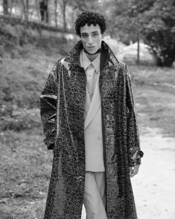 Monochrome portrait of a man in an animal-print trench coat posing outdoors in a park with a background of trees and foliage.