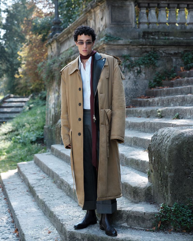 A stylish adult male stands on stone steps outdoors wearing a long tan coat, dark trousers, and boots, with trees and foliage in the backdrop.