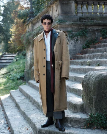 Model Walid Fiher poses on outdoor stone steps wearing an oversized camel trench coat, rectangular sunglasses, and black boots.