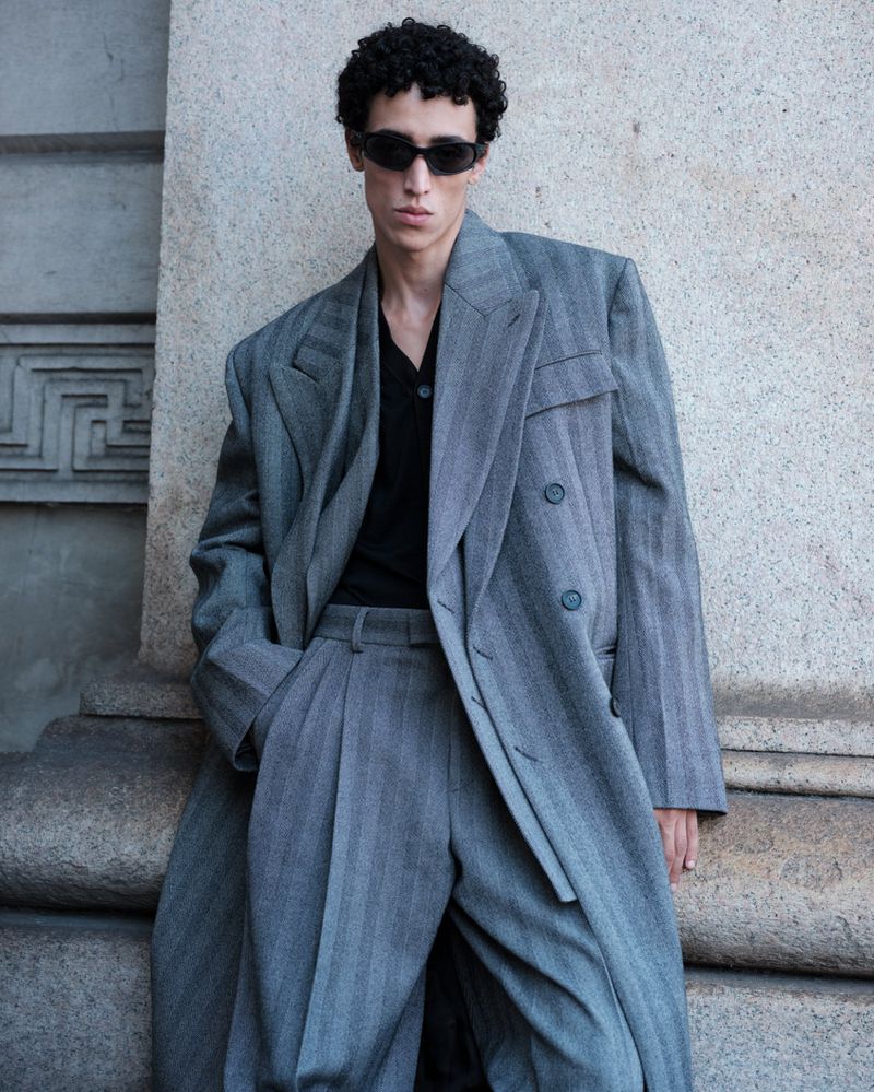 A cool-toned, medium shot of a male model with sunglasses and curly hair wearing a gray pinstripe suit and coat against a concrete wall.
