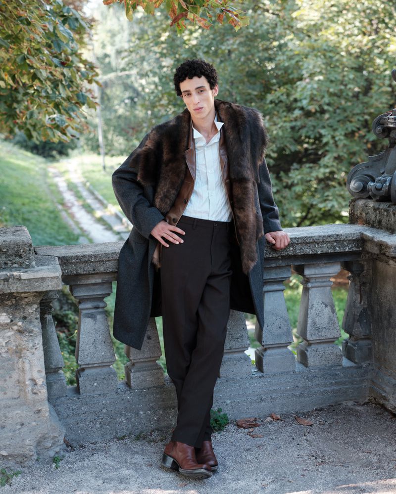A man in a fur-collared coat poses against a stone wall in a park, wearing brown pants and boots under the shade of green foliage.