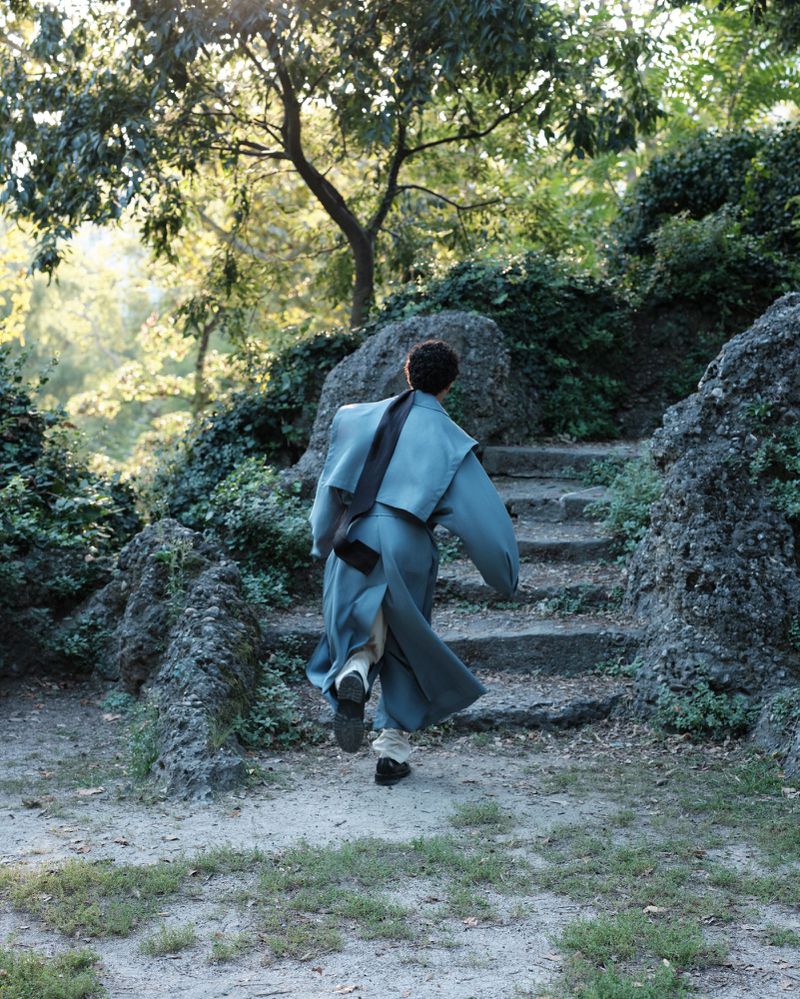 Back view of a person ascending stone steps in a park, wearing a long coat and carrying a bag, surrounded by lush greenery.
