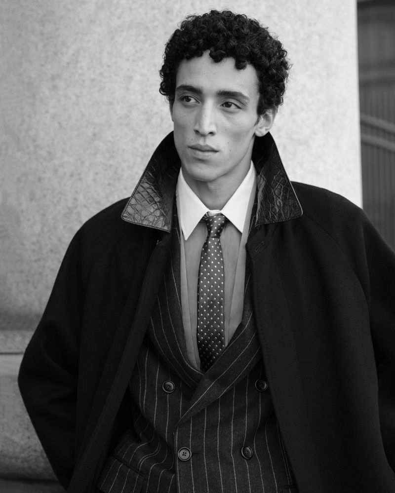 Black and white portrait of an adult male model wearing a suit and a coat, looking to the side in front of a neutral background.