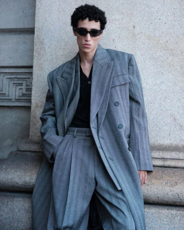Model Walid Fiher wearing an oversized grey wool overcoat, pleated suit trousers, and black sunglasses against a stone building.