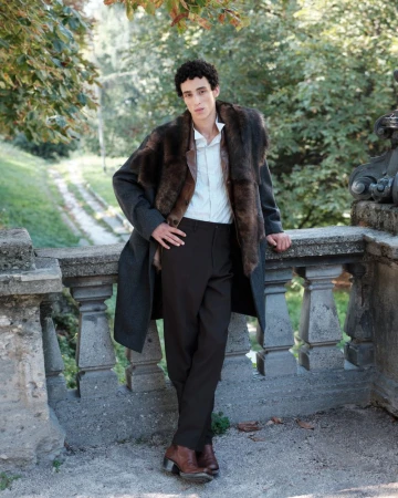 A male model leaning against a stone balustrade wearing a furry fur collar coat, white shirt, and dark trousers outdoors.