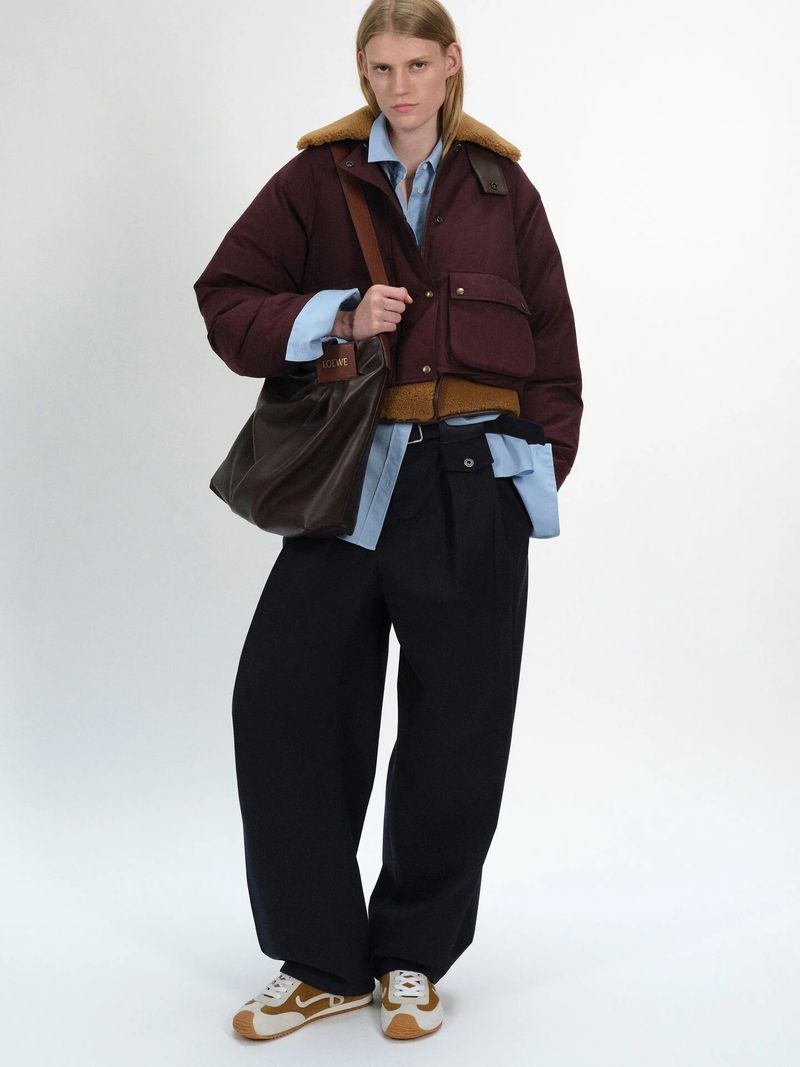 A fair-skinned woman poses in an oversized maroon puffer jacket, light blue shirt, black pants, and vintage running shoes on a white backdrop.