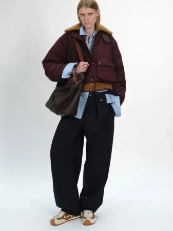 Model wearing a burgundy puffer jacket with a brown collar, light blue shirt, dark trousers, and carrying a brown leather shoulder bag.