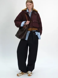 Model wearing a burgundy puffer jacket with a brown collar, light blue shirt, dark trousers, and carrying a brown leather shoulder bag.
