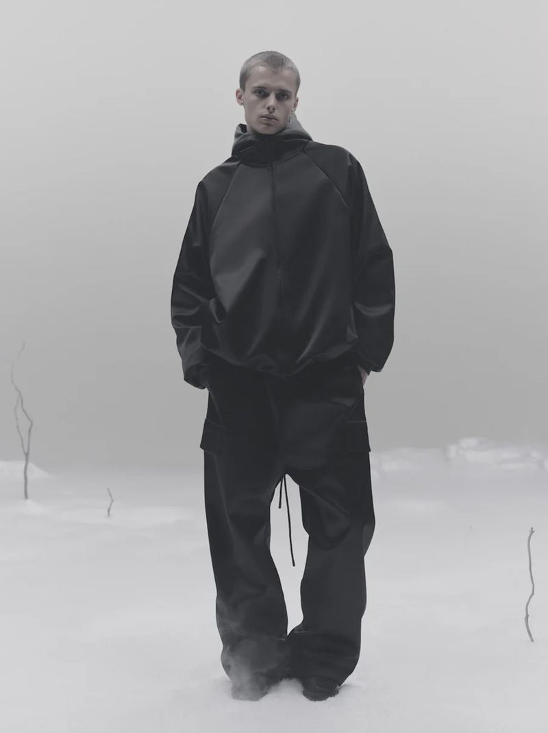 Man in black hooded sweatshirt and baggy pants standing in stark landscape. Fashion shot with minimalist aesthetic.