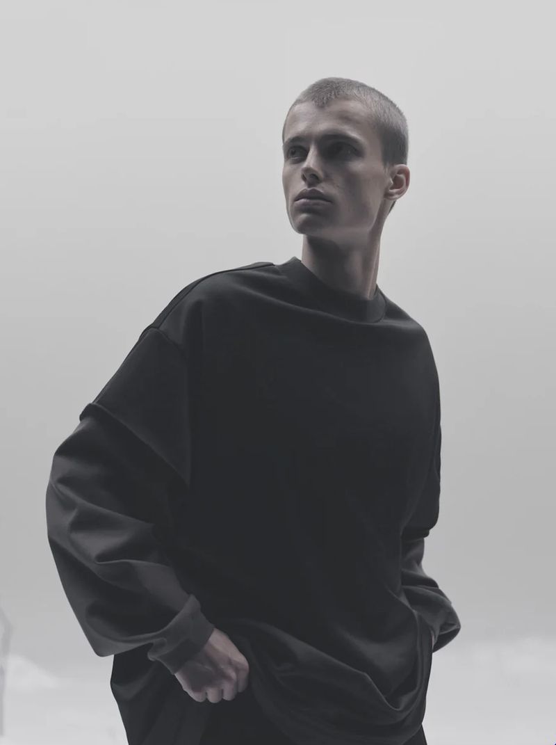 Portrait of a young adult male looking away, wearing a dark, oversized sweatshirt against a bright, neutral background in a studio setting.
