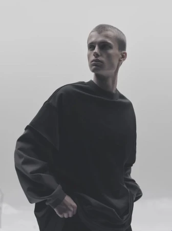 Portrait of a young adult male looking away, wearing a dark, oversized sweatshirt against a bright, neutral background in a studio setting.