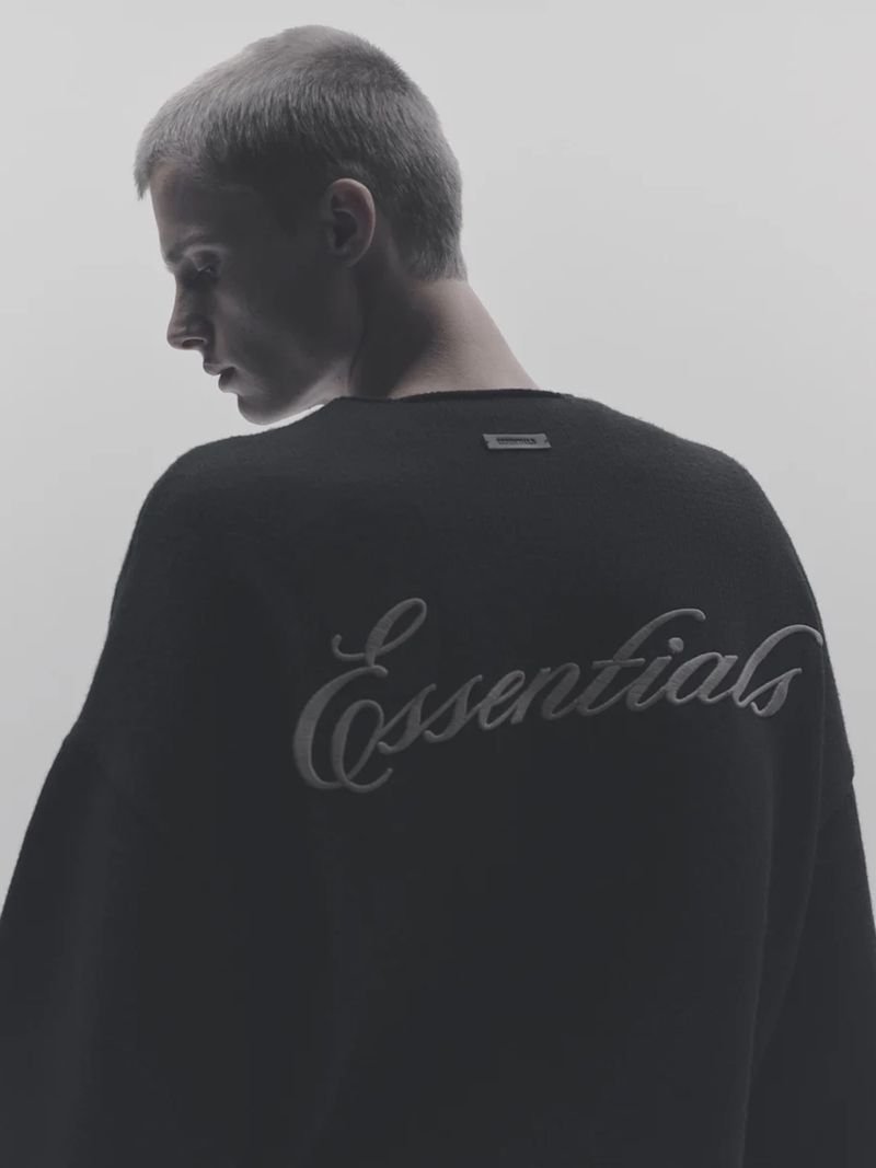 Back view of a light-skinned person with a shaved head wearing a black sweater with the word 'Essentials' embroidered on the back.