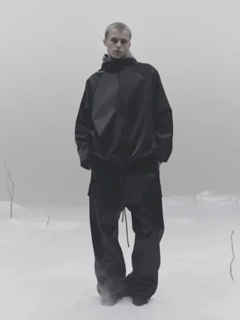 A young man wearing a dark hooded sweatshirt and trousers standing outdoors against a misty grey background.