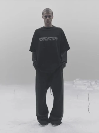 A model standing outdoors in a snowy landscape wearing a dark t-shirt and matching trousers for Fear of God Essentials Winter 2025.