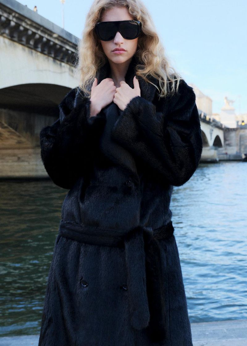 A woman with blond curly hair and black sunglasses and coat poses near a river and bridge in an outdoor setting on a sunny day.