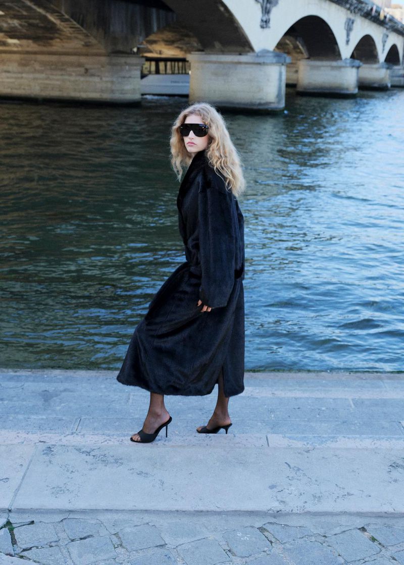 Model wearing a black coat and sunglasses by a river with a stone bridge in Paris for Mango