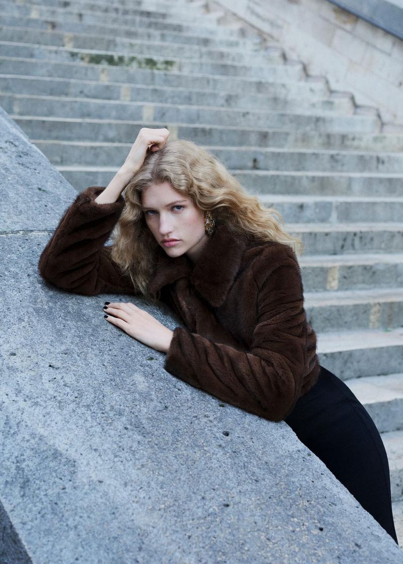 A woman with curly blonde hair wearing a fur coat and a black skirt, poses near concrete steps in an urban environment.