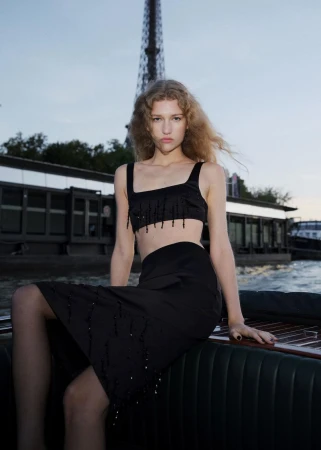 A blonde model poses in a black two-piece outfit on a boat with the Eiffel Tower in the background during daytime, fashion shoot.