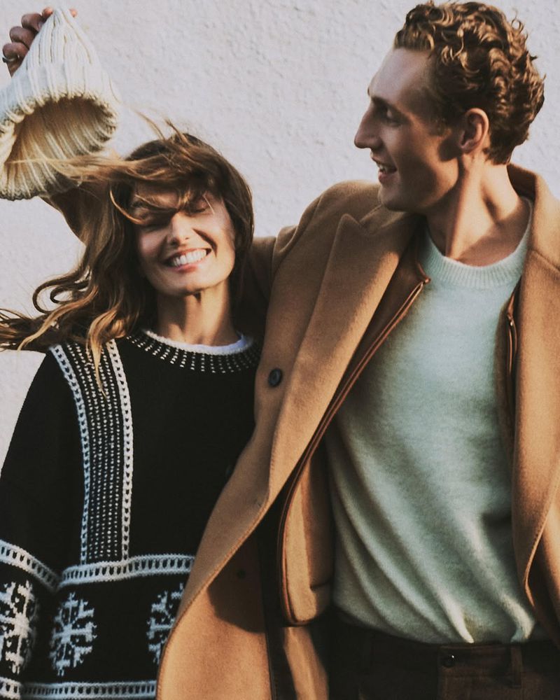 Smiling couple in winter fashion: woman in black patterned sweater, man in tan coat. Woman holds white knit hat near white background.
