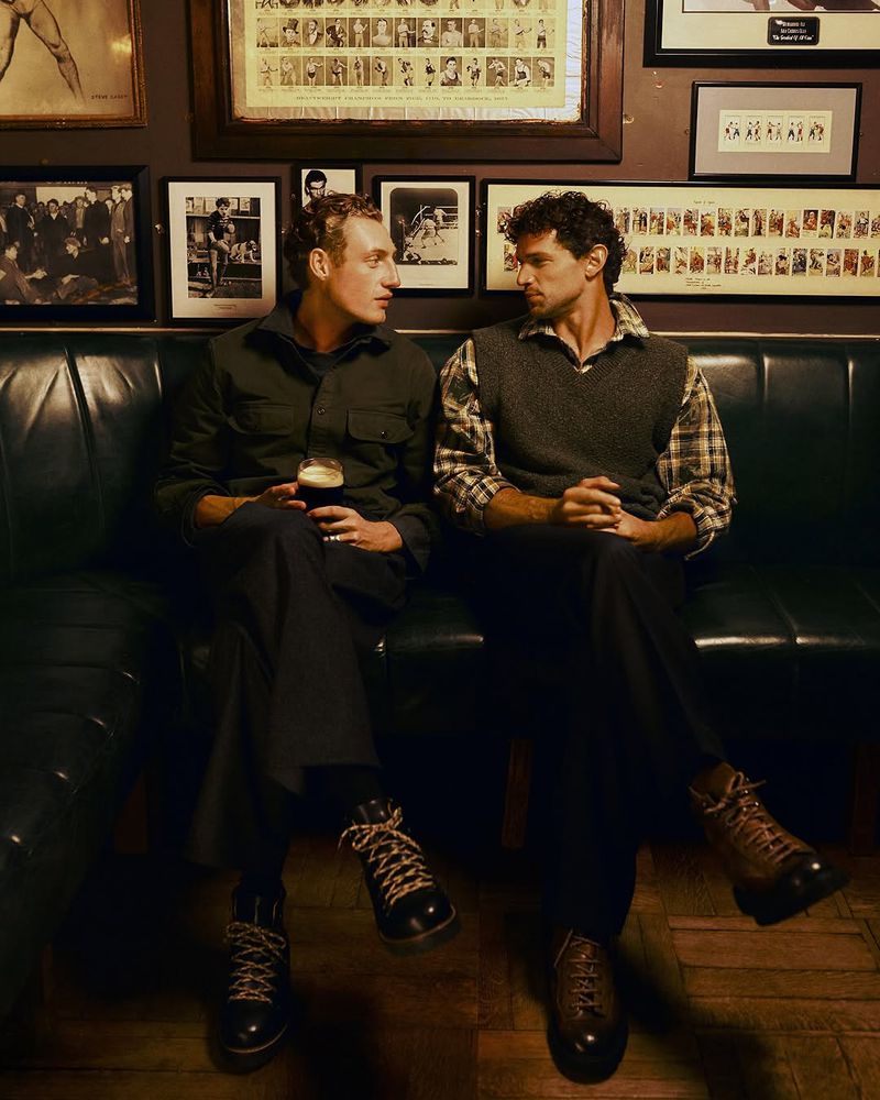Two men sit on a black leather bench in a dimly lit pub or bar with framed pictures on the wall. They are wearing casual clothing.