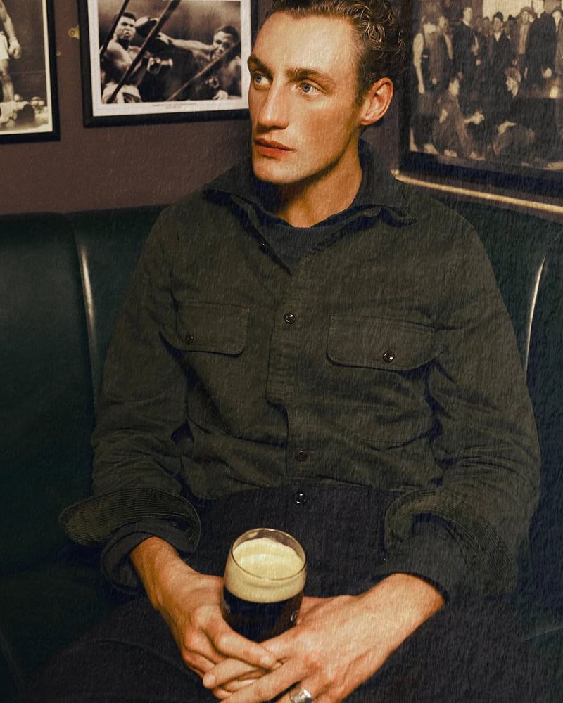 Man in a dimly lit bar holds a dark beer. He wears a button-down shirt and sits on a dark green banquette with boxing photos behind him.