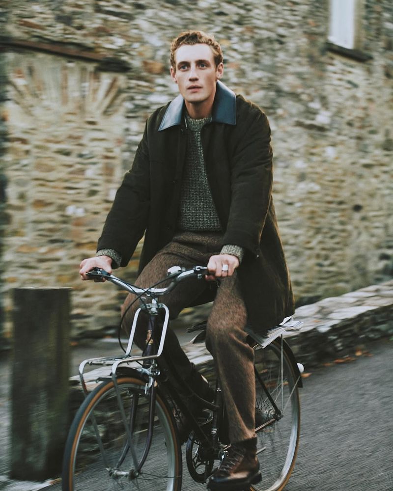 Man riding bicycle outdoors on a road near a stone building, wearing a dark coat, brown trousers and boots, travel and lifestyle photography.