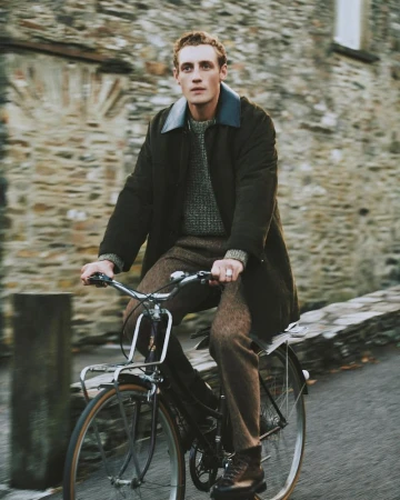 Man riding bicycle outdoors on a road near a stone building, wearing a dark coat, brown trousers and boots, travel and lifestyle photography.
