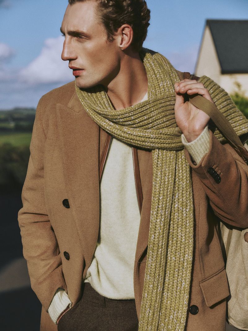 An adult male is captured in a medium shot wearing a tan overcoat, knit scarf, and brown trousers against an outdoor backdrop.