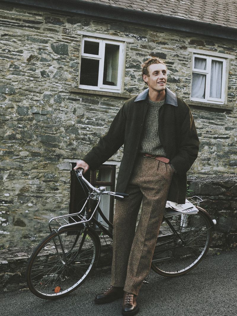 Man with tweed pants poses with bicycle against a stone house backdrop, wearing a jacket and sweater in an outdoor setting.