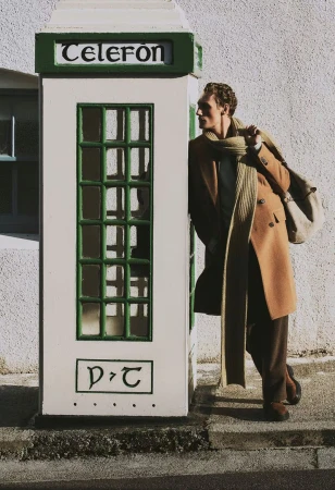Man in overcoat poses next to vintage phone booth with travel bag on city street. Green, white, and tan color palette.