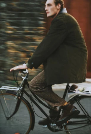 Adult male on bicycle with coat and tweed pants rides along blurred brick building, natural light, medium shot, outdoor, lifestyle.