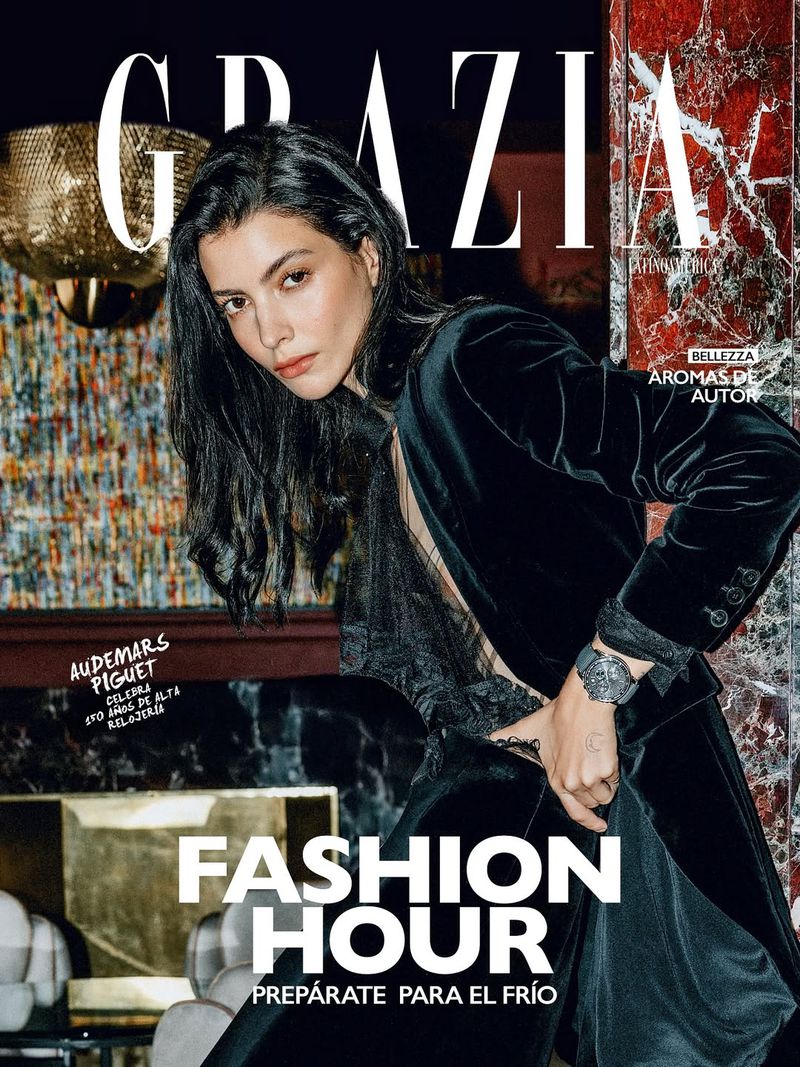Magazine cover featuring a dark-haired woman in a black velvet blazer and sheer top, glancing at the viewer with golden lighting and marble backdrop.