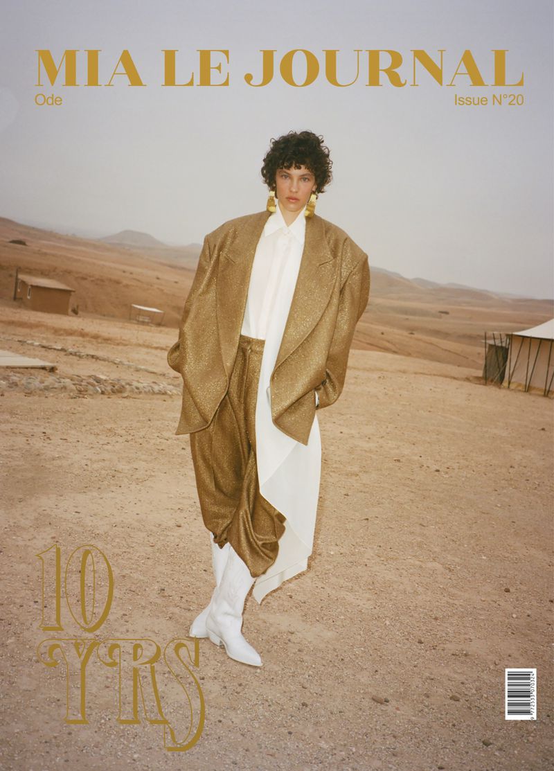 Magazine cover featuring a fashion model in a desert landscape wearing a gold coat, white pants, and white boots, promoting 'Mia Le Journal'.